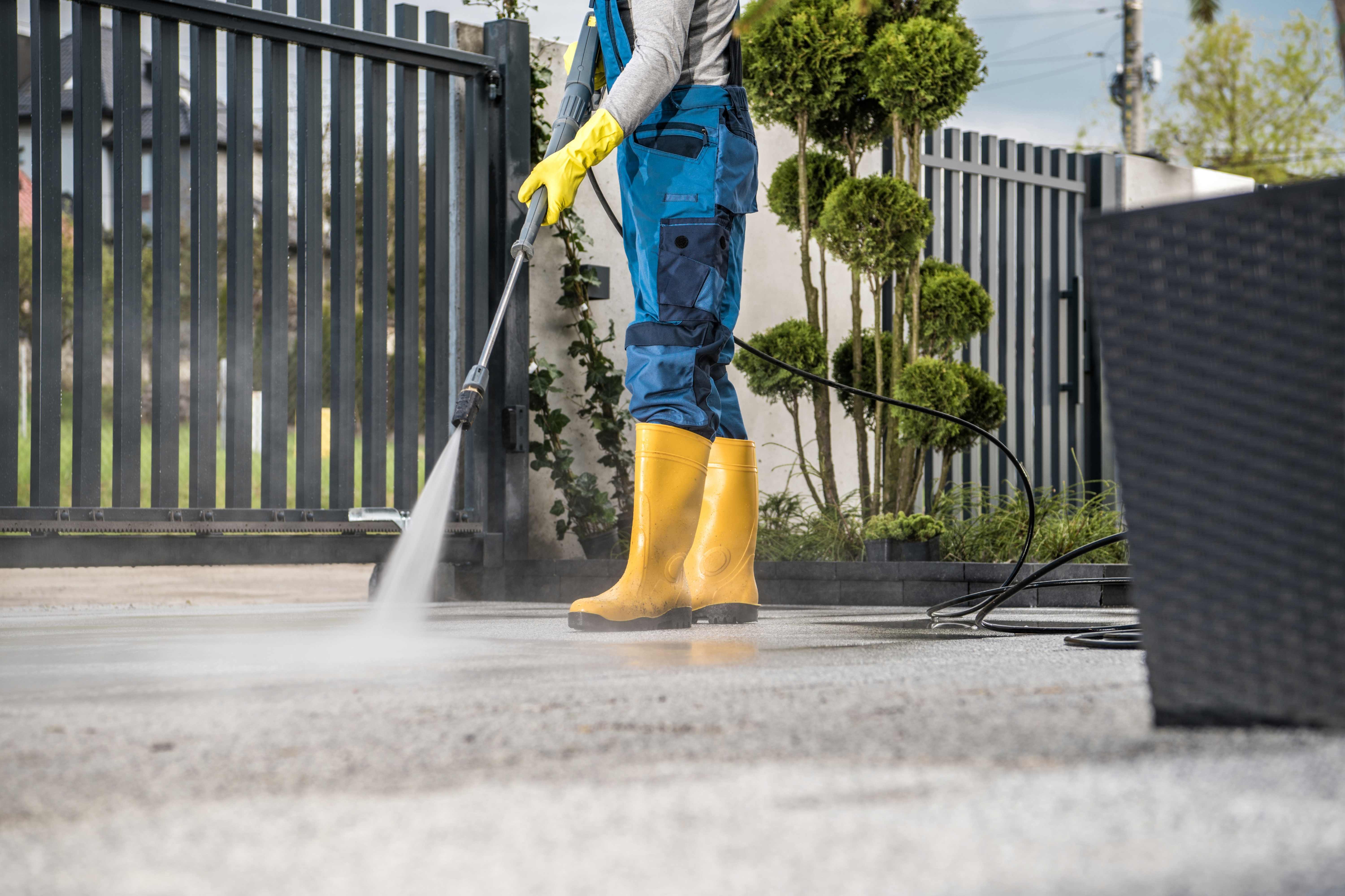 Professional worker in blue coveralls and yellow protective boots performing pressure washing driveway cleaning service in Phoenix